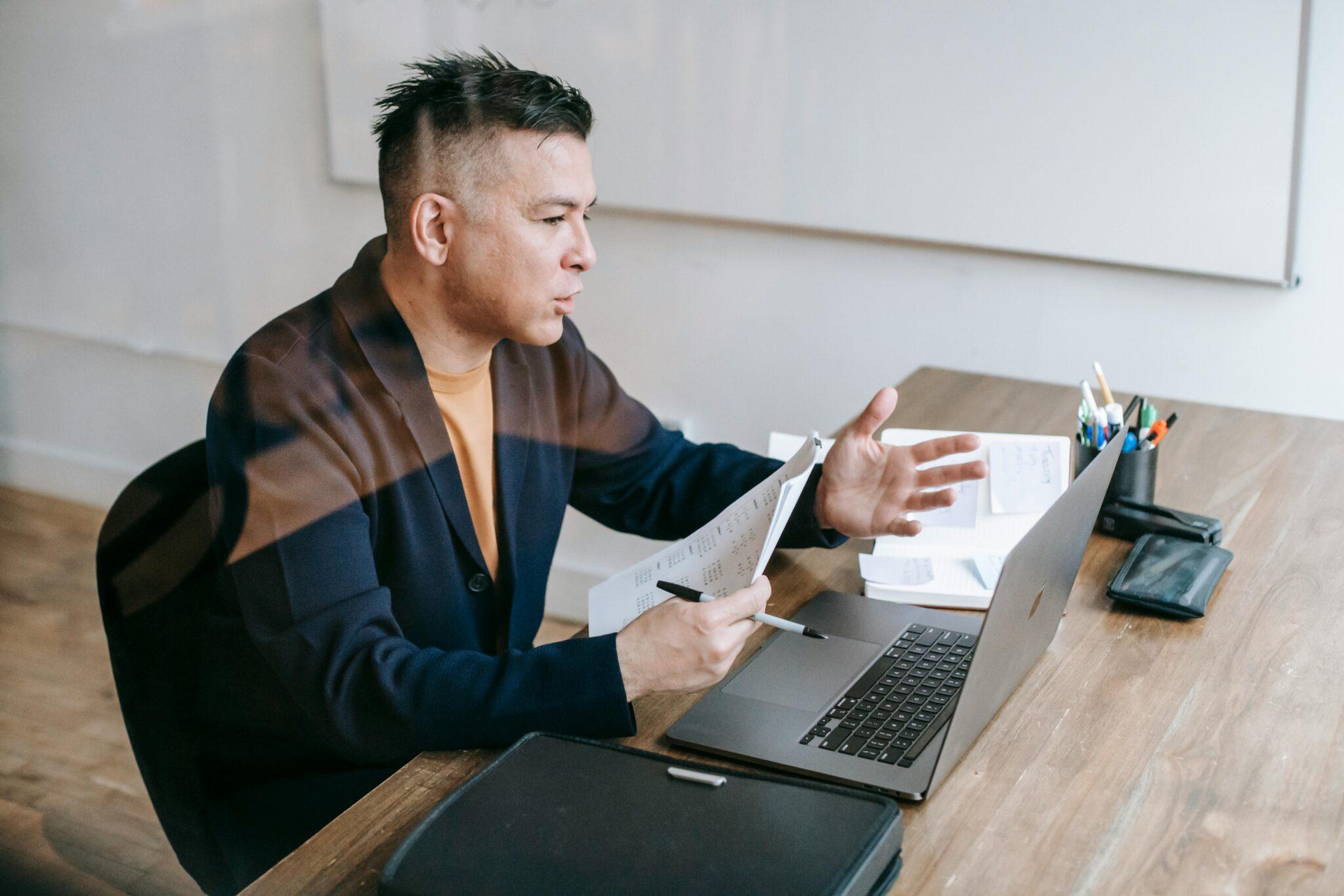 man talking to an open laptop holding papers